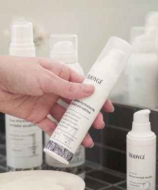 une main attrape la crème hydratante Hodygé dans une salle de bain